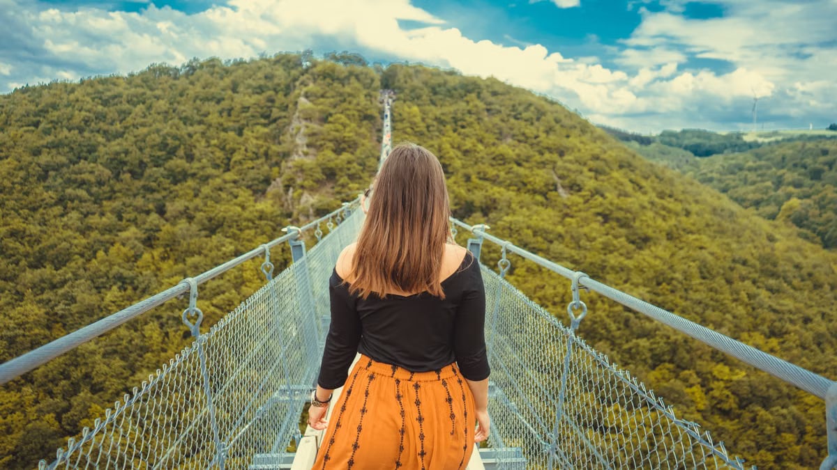 A woman walks along a canopy bridge set amidst a lush green forest and scenic hills.